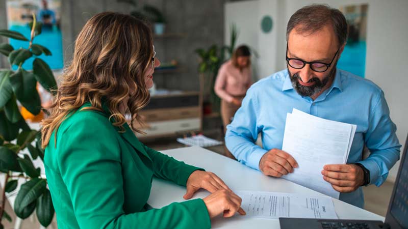 Homem adulto estrangeiro a ser atentido num balcão de um banco por uma senhora com casaco verde; vêem uma série de documentos no balcão